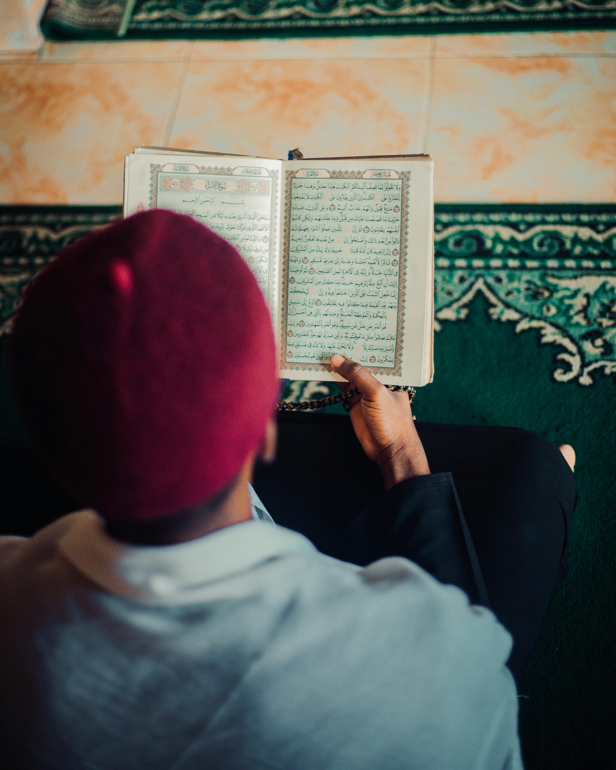 Man in prayer cap reads Quran in a serene mosque setting, focused on devotion.